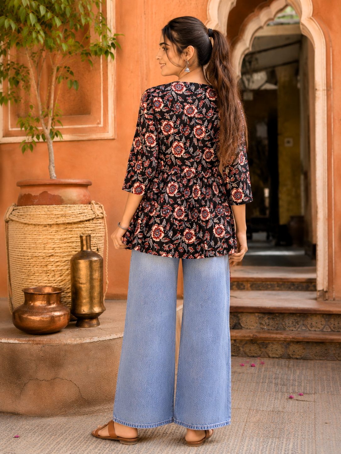 A woman is shown from behind wearing a black floral peplum top and wide-leg light blue jeans, standing in front of a warm terracotta-colored building with decorative vases.