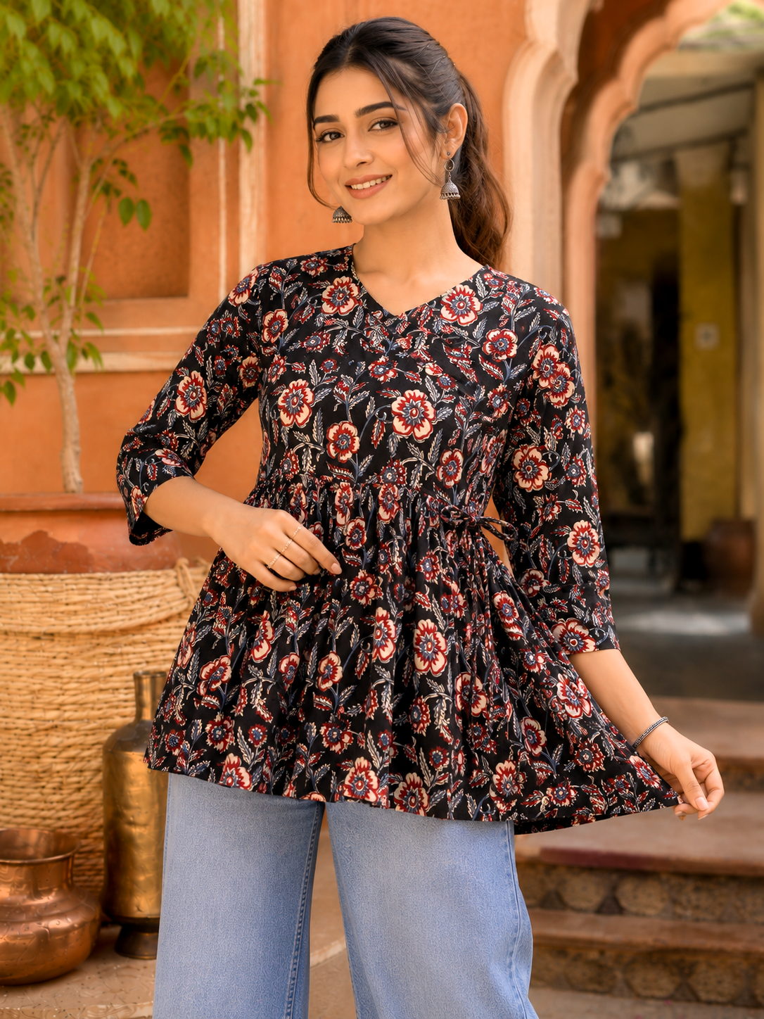 A woman wearing a black floral peplum top with three-quarter sleeves and light blue jeans, standing in front of a warm-toned architectural background with arches and plants.