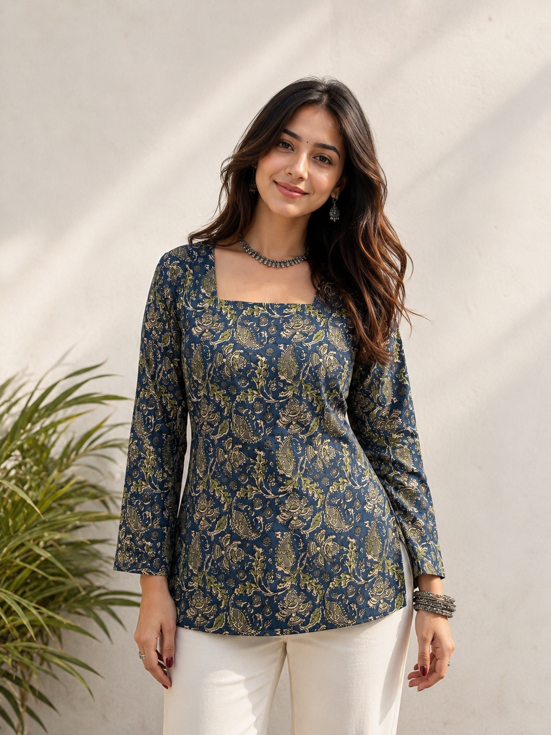A woman modeling a navy blue long-sleeve kurta with a gold floral print, paired with white pants, standing against a textured light beige wall with soft shadows.