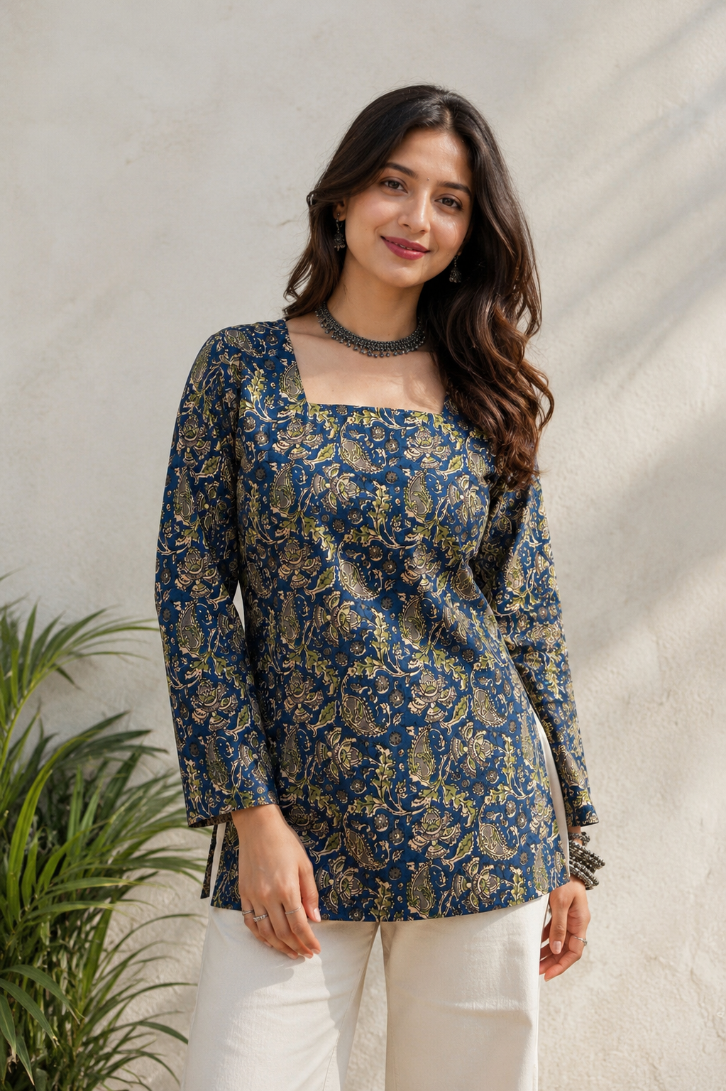 A woman wearing a navy blue long-sleeve kurta with a gold floral block print, paired with white trousers, standing against a textured light beige wall with a green plant on the left.