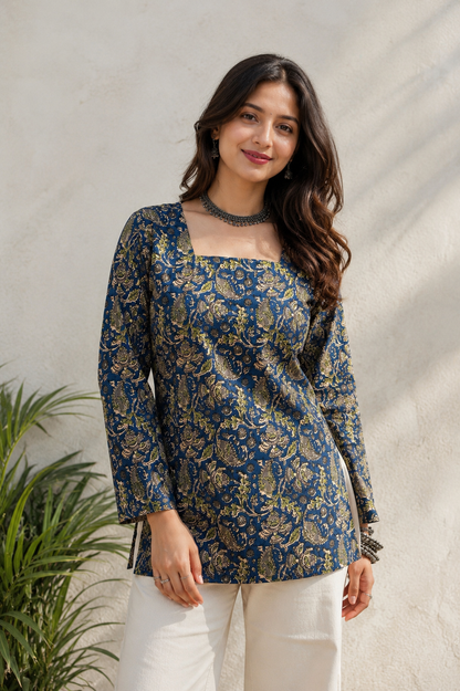 A woman wearing a navy blue long-sleeve kurta with a gold floral block print, paired with white trousers, standing against a textured light beige wall with a green plant on the left.