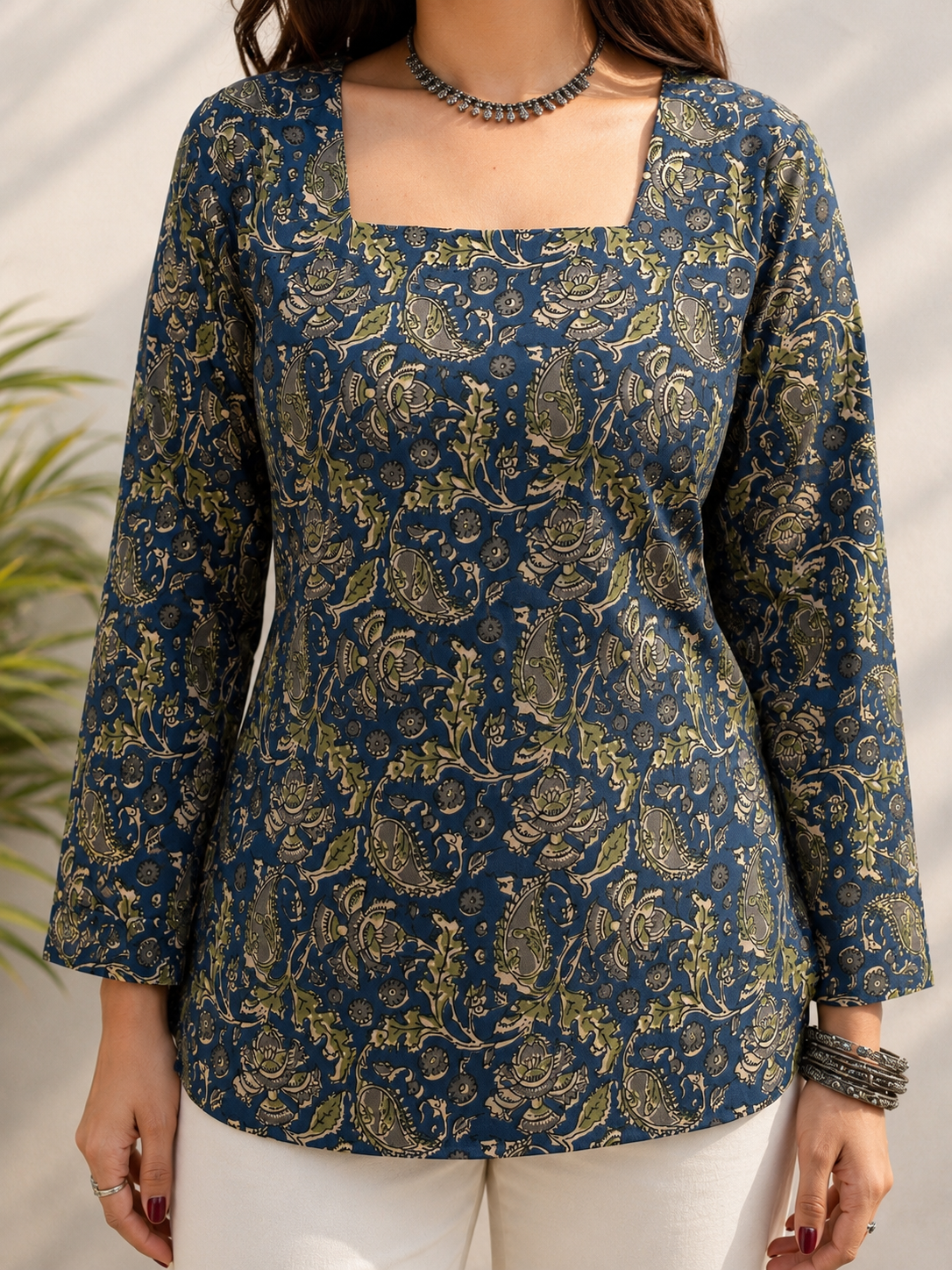 A woman wearing a navy blue long-sleeve top with a gold floral block print pattern, paired with white pants. The top features a square neckline and an all-over ornate paisley and floral design.