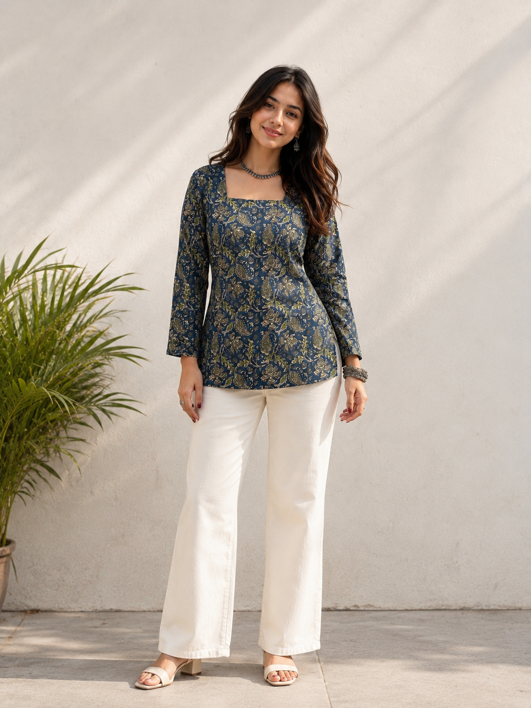 A woman modeling a navy blue floral long-sleeve top with a square neckline, paired with white wide-leg pants and beige sandals, standing against a textured light gray wall with a potted plant on the left.