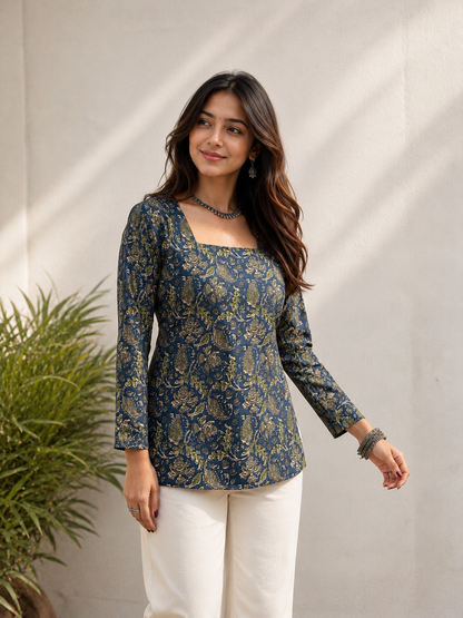 A woman modeling a navy blue long-sleeve kurta with a gold floral pattern, paired with white pants, standing against a neutral beige wall with soft shadows.