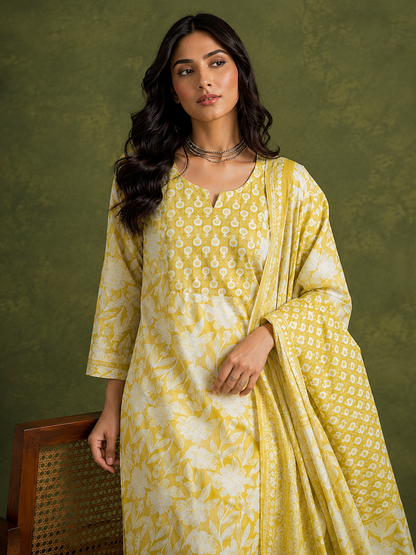A woman models a yellow and white printed kurta set with a matching dupatta against a textured olive green studio backdrop.